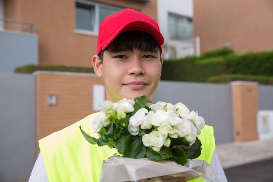 A young delivery person wearing a red cap and high-visibility vest is standing outside a modern residential building, holding a bouquet of fresh white flowers wrapped in white paper with dark green le