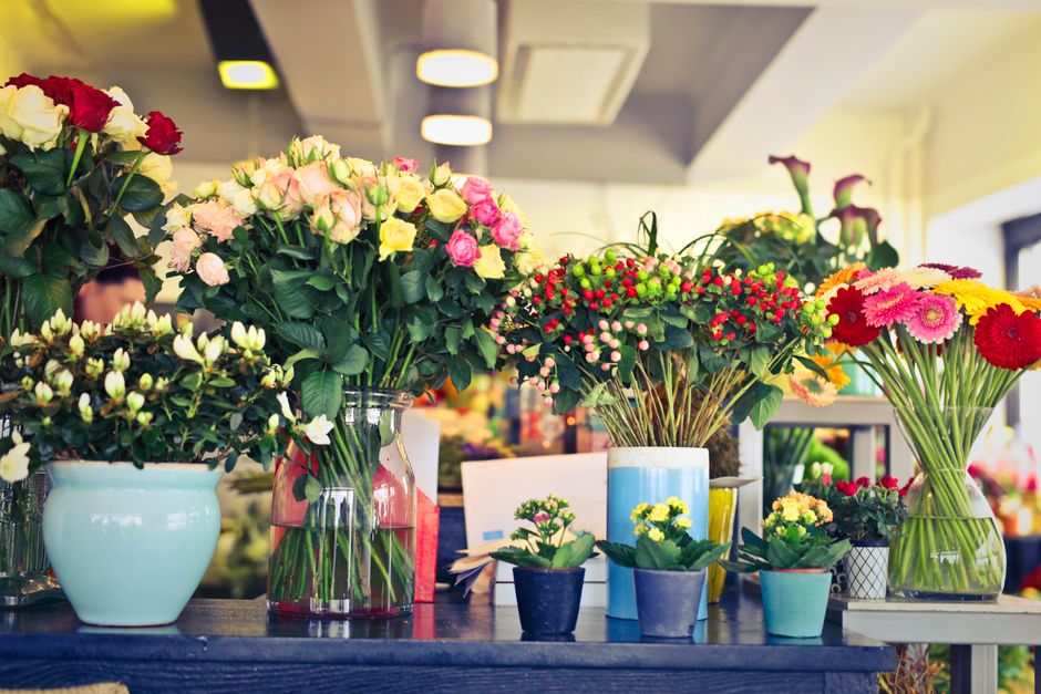 A vibrant display of fresh floral arrangements on a dark wooden table inside a well-lit flower shop, featuring bouquets of pink, white, yellow, and red roses, gerbera daisies, and small flowering plan