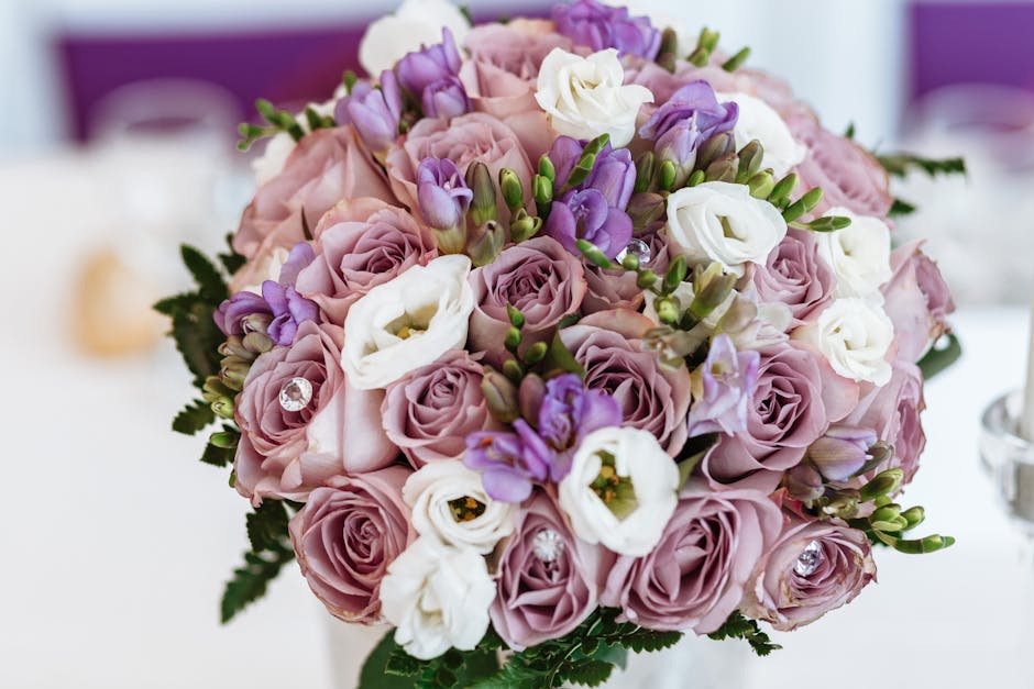 A close-up of a floral bouquet featuring light mauve roses, white eustomas, and purple freesia, arranged in a compact, rounded style. The flowers are fresh with soft, velvety petals and visible green 