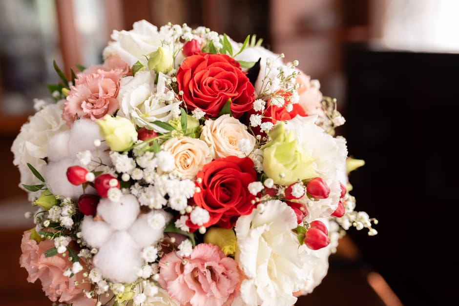 A close-up of a floral bouquet featuring vibrant red roses, soft peach roses, and delicate white lilies, complemented by pink and white spray roses, baby's breath, and greenery, all arranged in a lush