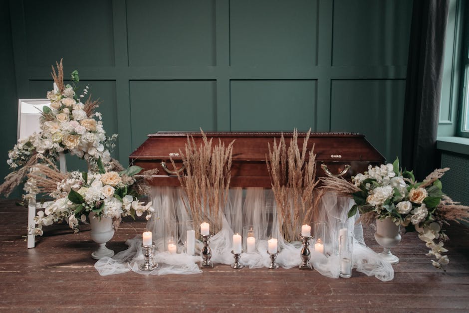 A wooden casket is positioned against a dark green wall, decorated with elegant floral arrangements on either side. The arrangements feature white roses, hydrangeas, and lilies, complemented by beige 