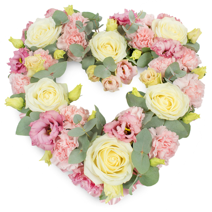 This image shows a meticulously crafted heart-shaped funeral wreath, its pastel blooms and cool foliage arranged so densely that the entire form appears plush and cushion-like. The heart is perfectly defined, with smooth, rounded sides and a gentle dip at the top, all traced in soft florals that glow against a pristine white background. Dominating the composition are large cream and pale yellow roses, their velvety petals curled in tight yet generous spirals, creating a sense of serenity and steadfast love. These roses sit at balanced intervals around the heart, acting as visual anchors in much the same way familiar landmarks anchor the streets of Mile End. Between them, frothy clusters of light pink carnations spill outward, their finely frilled petals forming layers of gentle texture that soften the overall look, like a comforting blanket. Deeper rose-pink to mauve lisianthus blooms are scattered thoughtfully throughout, their more open, bell-like forms and subtle shadows adding depth and emotional richness to the design. Small yellow-green buds, some barely opening, peep out from the arrangement, hinting at renewal and the continuity of life. Interwoven among the blooms are slender stems of silvery-green eucalyptus, their rounded leaves catching the light and providing a cool, calm contrast that frames the heart and gives it structure. The colours remain restrained yet uplifting: blush pinks, warm creams, soft yellows, and muted greens working together to evoke tenderness, remembrance, and quiet respect. Crafted with the sensitivity of an experienced East London florist, this heart-shaped wreath would feel very much at home at a funeral or memorial service near Mile End, offering a graceful, heartfelt tribute to a life dearly cherished.